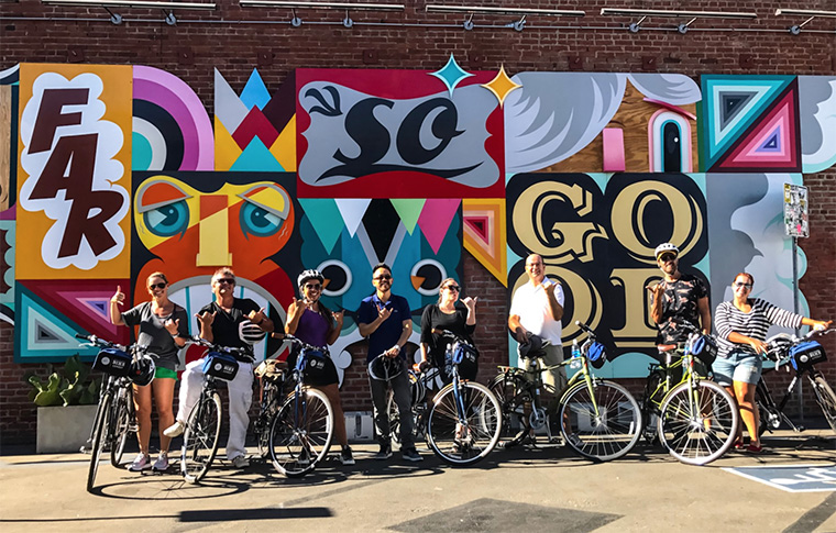 Group of tour goers and guide posing with their bikes and helmets in front of a mural